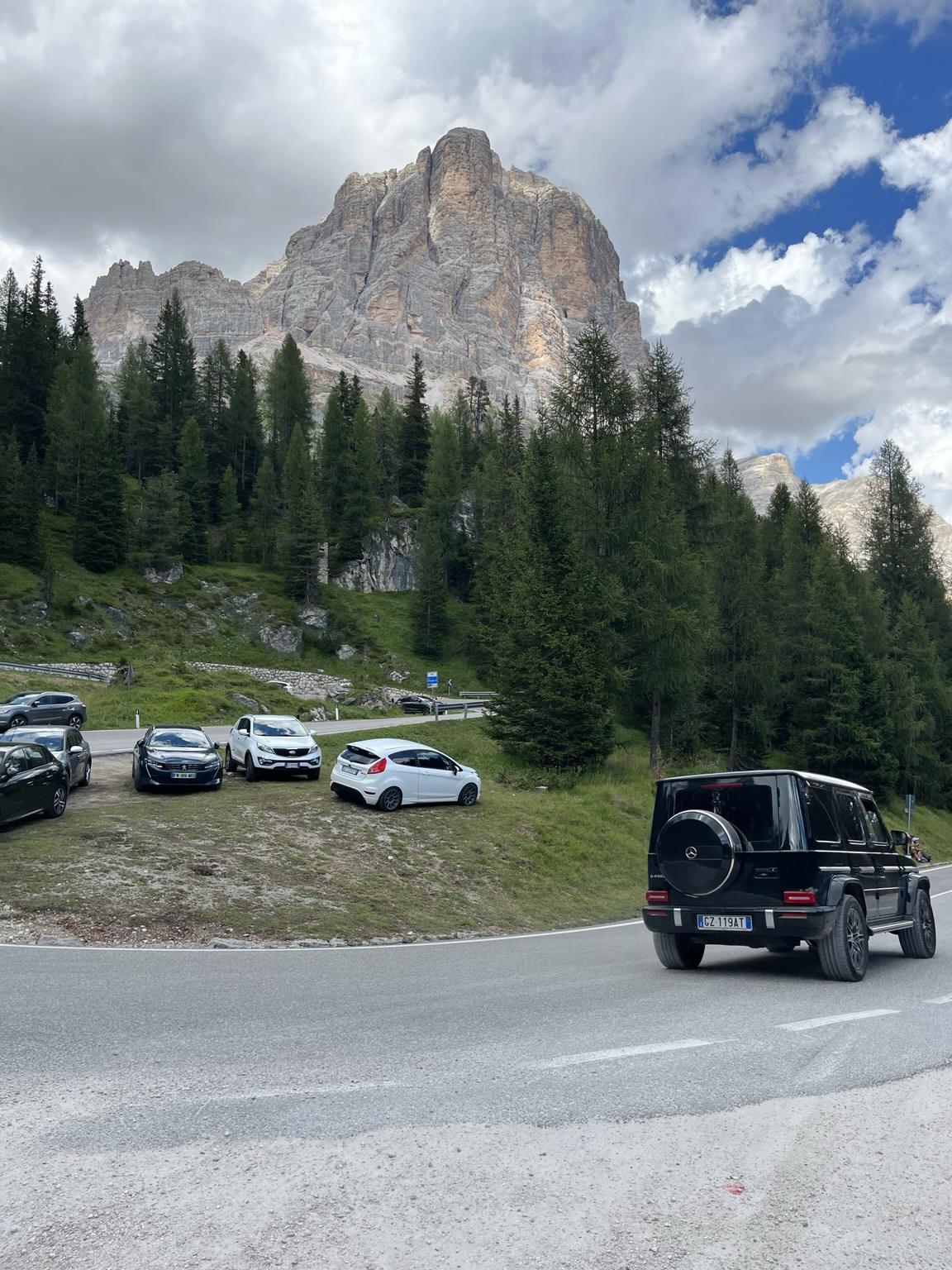 Cars parked on the grassy patch within a hairpin turn, a Mercedes G Klasse zooms past in the foreground